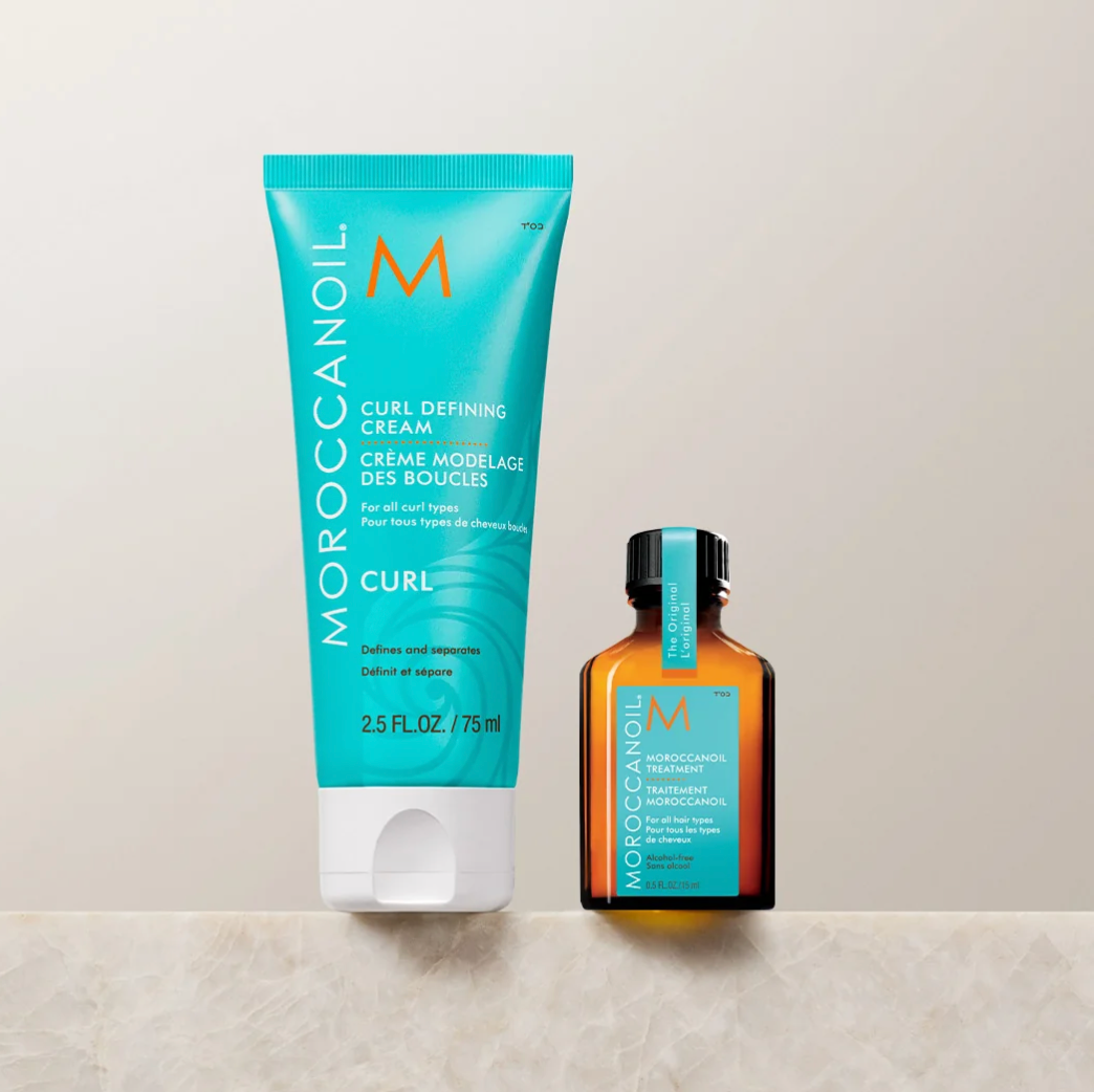 Moroccanoil Curl Defining Cream in a turquoise squeeze tube and a matching Moroccanoil Hydrocarbon Treatment in an amber glass bottle, displayed on a light beige marble surface against a neutral beige background.