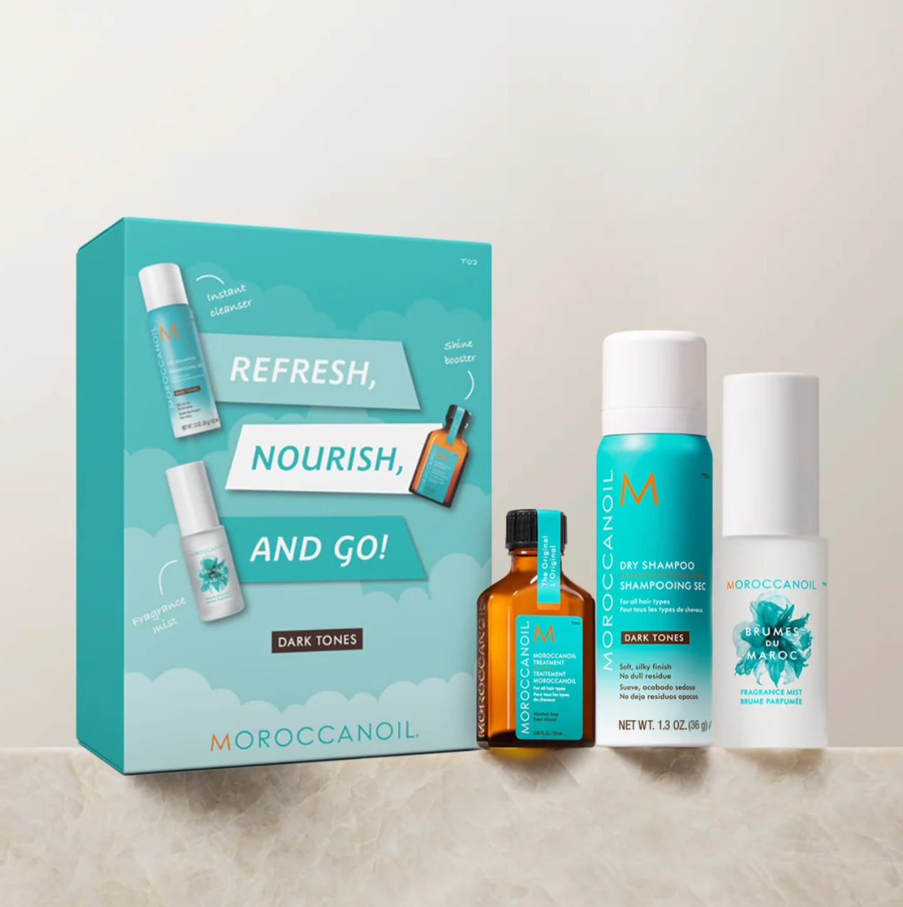 Moroccanoil 'Refresh, Nourish, and Go!' Dark Tones hair care set, featuring a teal gift box, a brown amber bottle, a blue canister, and a white bottle, arranged on a light beige marble surface against a neutral background.