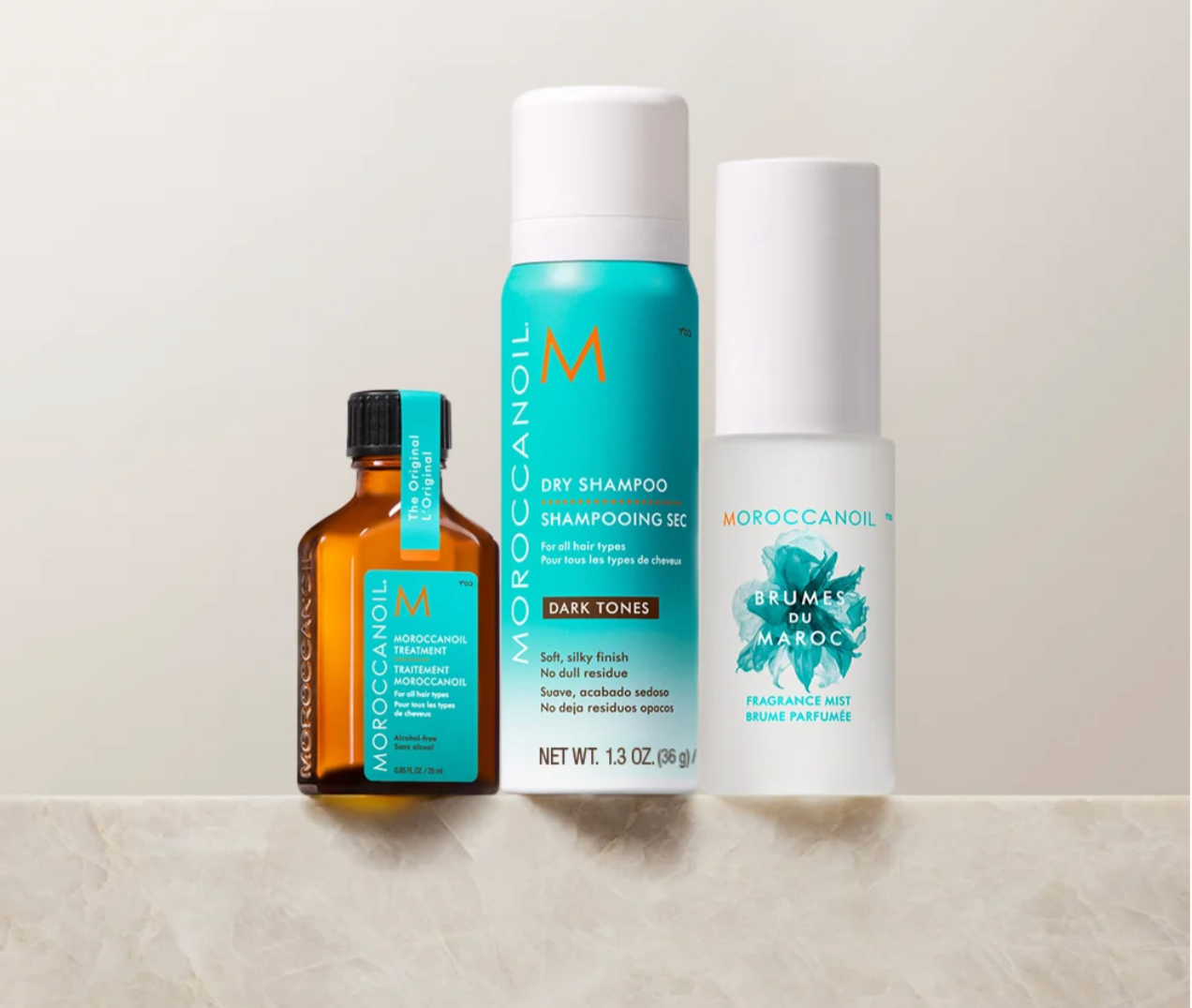 Three Moroccanoil haircare products arranged on a light stone surface against a warm off-white background: a dark amber bottle with a black cap, a turquoise aerosol can, and a white cylindrical bottle with blue floral graphics.