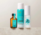 Three Moroccanoil haircare products arranged on a light stone surface against a warm off-white background: a dark amber bottle with a black cap, a turquoise aerosol can, and a white cylindrical bottle with blue floral graphics.
