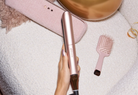 A hand holds a rose gold ghd hair straightener over a textured white blanket, with a matching pink hairbrush and a pink pouch nearby.